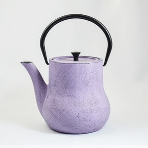 May include: A lavender purple cast iron teapot with a black handle and lid. The teapot has a textured surface.