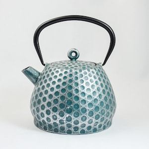 Op de afbeelding: Een tealblauwe gietijzeren theepot met een zwarte handgreep en een getextureerd, gebollet oppervlak.