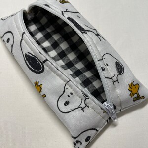 Pu&ograve; includere: Un astuccio per matite bianco con cerniera e fodera a scacchi bianco e nero. L'esterno presenta un motivo ripetitivo di cani di cartoni animati in bianco e nero e uccelli gialli.