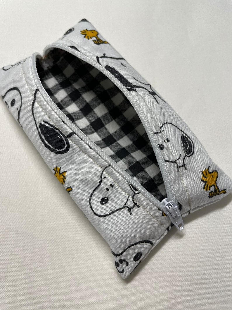 Pu&ograve; includere: Un astuccio per matite bianco con cerniera e fodera a scacchi bianco e nero. L'esterno presenta un motivo ripetitivo di cani di cartoni animati in bianco e nero e uccelli gialli.