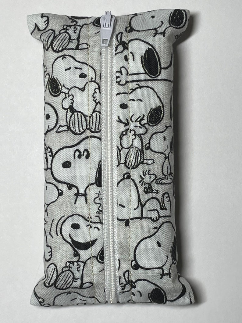 Op de afbeelding: Een witte ritszak met een zwart-witte print van Snoopy-figuren. De tas is gemaakt van stof en heeft een ritssluiting.