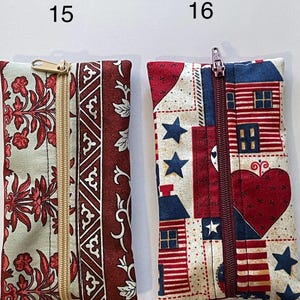 May include: Two rectangular fabric pouches with zips. Pouch 15 features a light green and dark red floral pattern with a gold zip. Pouch 16 displays a red, white, and blue patriotic print with houses, hearts, and stars, featuring a dark red zip.