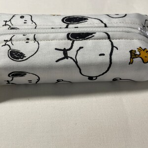 Pu&ograve; includere: Una pochette bianca con cerniera con una stampa di cartoni animati in bianco e nero di Snoopy e Woodstock. La pochette &egrave; realizzata in tessuto e ha una chiusura a cerniera.