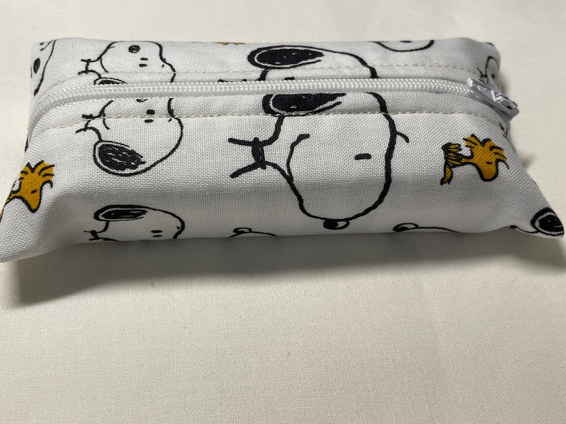 Pu&ograve; includere: Una pochette bianca con cerniera con una stampa di cartoni animati in bianco e nero di Snoopy e Woodstock. La pochette &egrave; realizzata in tessuto e ha una chiusura a cerniera.