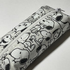 Op de afbeelding: Een wit potlood etui met een zwart-witte print van Snoopy en zijn vrienden. Het etui heeft een rits.