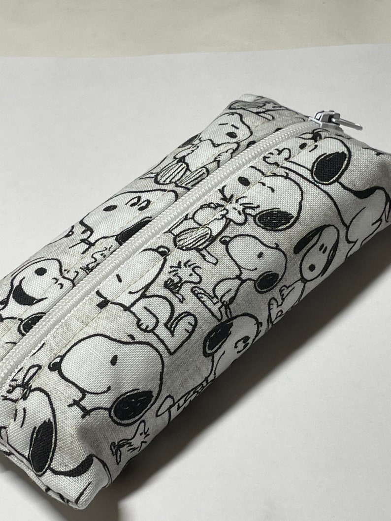 Op de afbeelding: Een wit potlood etui met een zwart-witte print van Snoopy en zijn vrienden. Het etui heeft een rits.