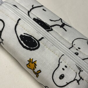Pu&ograve; includere: Borsa bianca con cerniera con una stampa in bianco e nero di Snoopy e Woodstock. La borsa &egrave; realizzata in tessuto e ha una cerniera bianca.