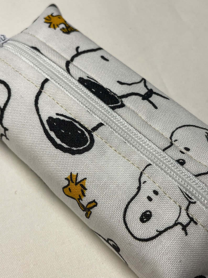 Pu&ograve; includere: Borsa bianca con cerniera con una stampa in bianco e nero di Snoopy e Woodstock. La borsa &egrave; realizzata in tessuto e ha una cerniera bianca.
