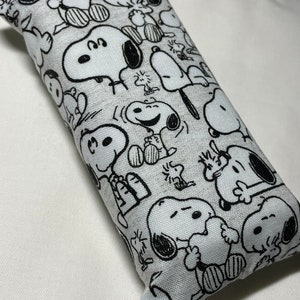 Op de afbeelding: Een wit en zwart gepatroneerd stoffen oogkussen met een herhalend ontwerp van Snoopy-personages. Het kussen is rechthoekig en heeft een zachte, pluche textuur.
