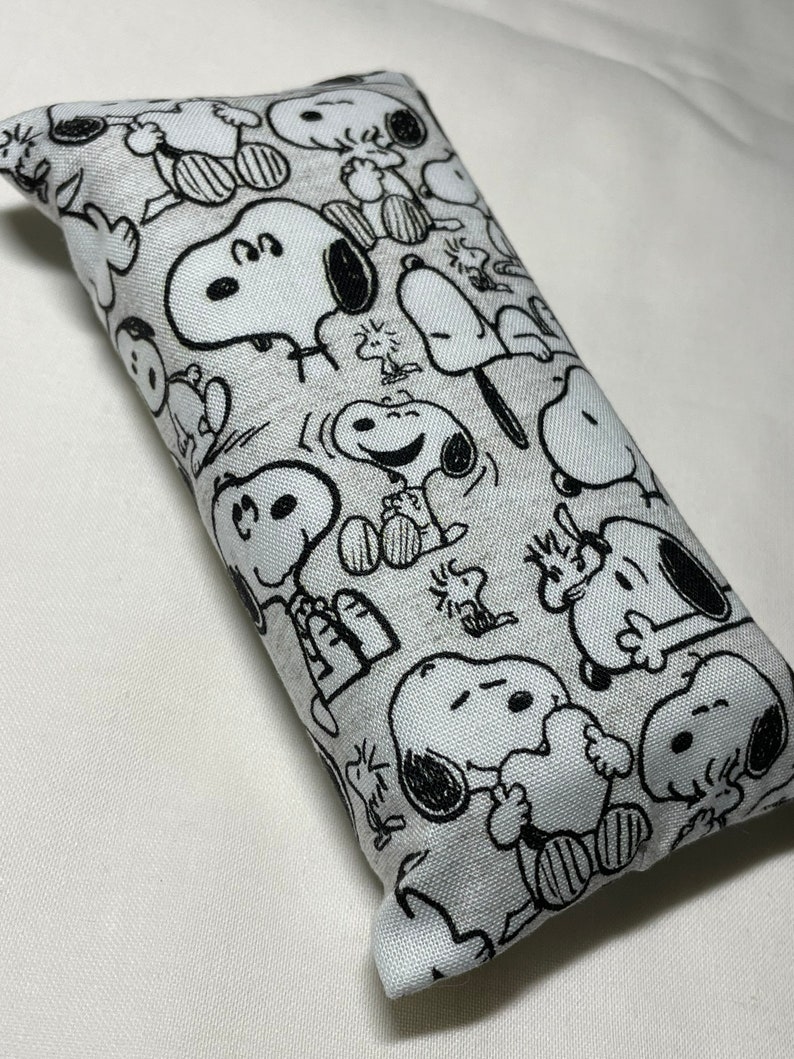 Op de afbeelding: Een wit en zwart gepatroneerd stoffen oogkussen met een herhalend ontwerp van Snoopy-personages. Het kussen is rechthoekig en heeft een zachte, pluche textuur.