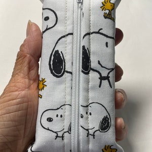 Pu&ograve; includere: Borsa bianca con cerniera con un motivo ripetuto di cani di cartoni animati in bianco e nero e uccelli gialli. La borsa &egrave; chiusa e tenuta in una mano.