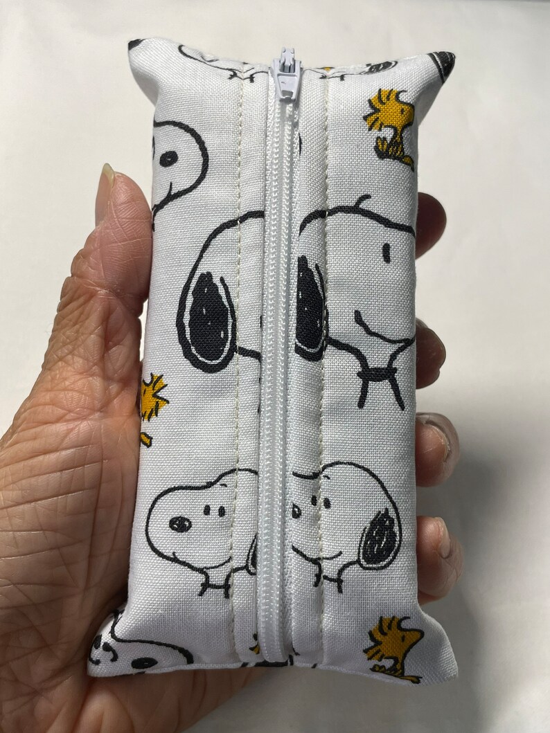 Pu&ograve; includere: Borsa bianca con cerniera con un motivo ripetuto di cani di cartoni animati in bianco e nero e uccelli gialli. La borsa &egrave; chiusa e tenuta in una mano.