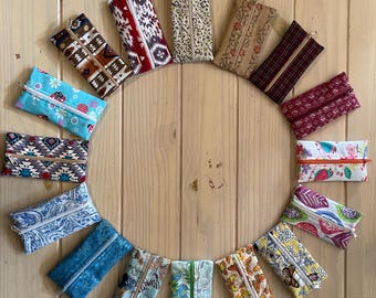 Op de afbeelding: Een cirkelvormige opstelling van vijftien rechthoekige stoffen etuis met ritssluiting, voorzien van diverse patronen zoals bloemen, geometrische vormen, paisley, vlinders en muzieknoten in uiteenlopende kleuren zoals blauw, rood, geel en bruin.