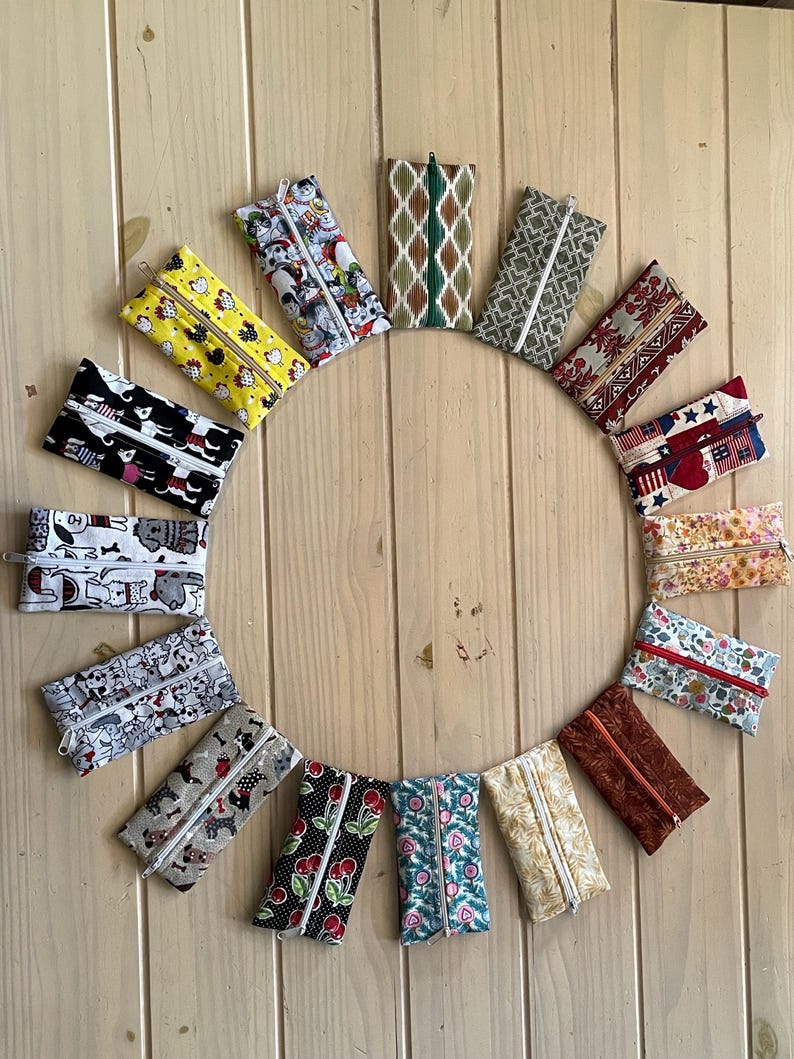 May include: A collection of fifteen rectangular fabric zip pouches arranged in a circle. Each pouch features a unique pattern, including floral, animal, geometric, and holiday-themed prints in various colours, all equipped with a central zip fastener.