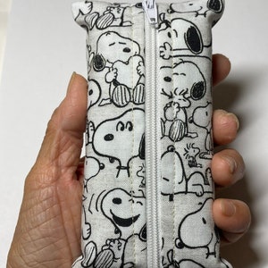 Op de afbeelding: Een witte ritszak met een zwart-witte print van Snoopy-figuren. De tas is gemaakt van stof en heeft een witte rits.