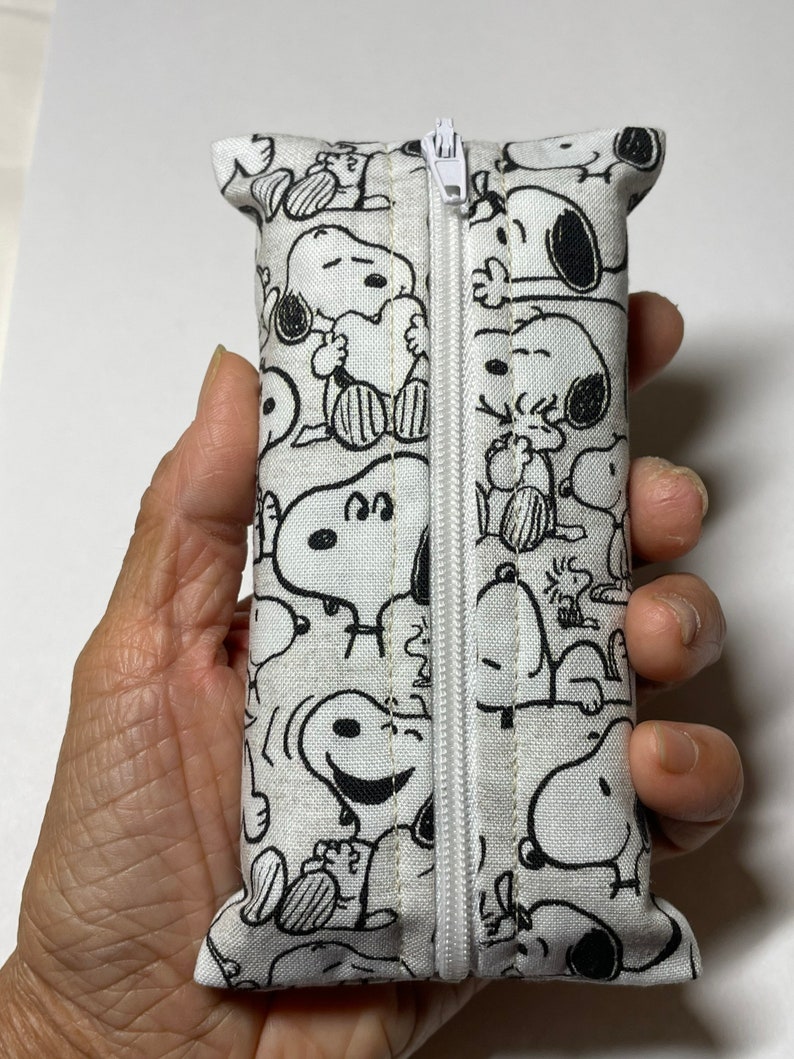 Op de afbeelding: Een witte ritszak met een zwart-witte print van Snoopy-figuren. De tas is gemaakt van stof en heeft een witte rits.