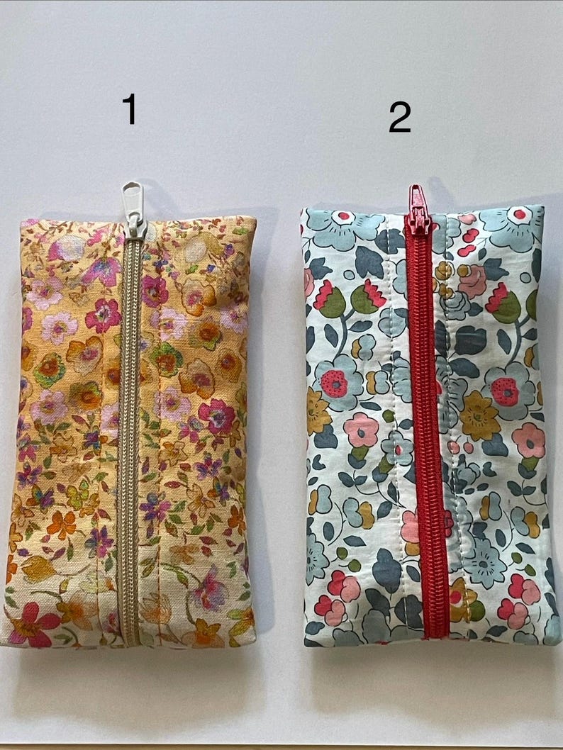 May include: Two rectangular fabric pouches with floral patterns. Pouch 1 features a yellow background with pink, orange, and green flowers and a silver zip. Pouch 2 has a light blue background with blue, pink, and yellow flowers and a red zip.