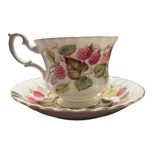 May include: A white teacup and saucer set with a floral design featuring pink raspberries and green leaves. The cup has a curved handle and gold trim. The saucer has a scalloped edge and a matching raspberry and leaf pattern.
