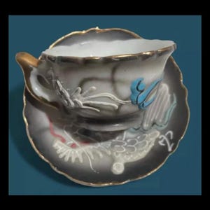 May include: A white teacup and saucer with a black and gold rim. The cup and saucer are decorated with a floral design and a blue bird. The saucer has a dragon design.