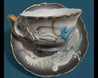 May include: A white teacup and saucer with a black and gold rim. The cup and saucer are decorated with a floral design and a blue bird. The saucer has a dragon design.