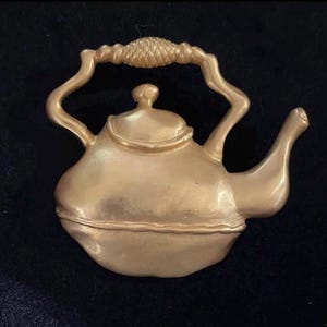 May include: A gold-colored teapot-shaped decorative object. The teapot has a curved handle with a textured design, a spout, and a lid with a small knob. The object is likely a brooch or a decorative element.