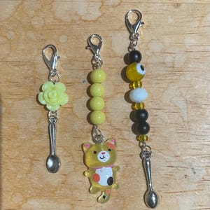 May include: Three handmade charms with silver-tone clasps. One charm features a yellow flower and a small spoon. Another has yellow beads and a cat charm. The third has black and yellow beads, a white bead, and a small spoon.