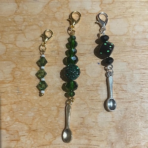 May include: Three decorative charms with lobster claw clasps. One charm features green faceted beads and white beads. Another has green beads and a small spoon charm. The third charm has black beads and a small spoon charm.