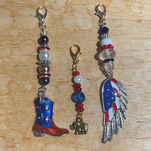May include: Three decorative charms with silver clasps. One features a red and blue cowboy boot, another a gold elephant, and the third a patriotic wing design. Each charm has beads in red, white, blue, and black.