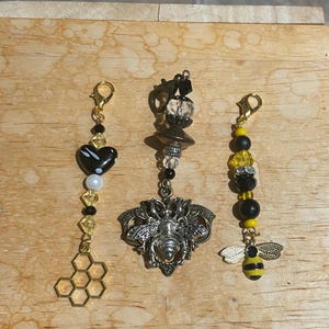 May include: Three handmade bee-themed charms. One charm features a black heart, a honeycomb, and gold beads. Another has a silver bee pendant and decorative beads. The third charm has a bee pendant and black and yellow beads.