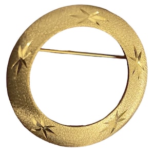 May include: A gold-toned circular brooch with a textured surface and four starburst designs evenly spaced around the outer edge. The brooch has a pin closure on the back.
