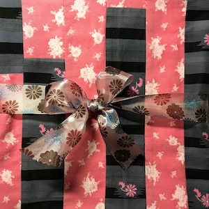 May include: A pink and gray fabric with a floral pattern is tied together with a large, patterned bow. The bow is made of a sheer, light pink fabric with brown and blue floral accents.