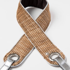 May include: A brown camera strap with silver trim and metal eyelets. The strap is made of a woven fabric and has a soft, padded lining.