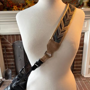 May include: A camera strap with a patterned design in shades of green, brown, and gold. The strap is attached to a black camera with a lens. The strap is displayed on a mannequin.