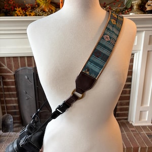 May include: A camera strap with a teal and gold patterned design. The strap is attached to a black camera with a lens. The strap is made of woven fabric and leather, with a metal ring and buckle.