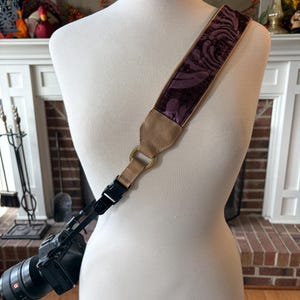 Może przedstawiać: Pasek do aparatu z kwiatowym wzorem w odcieniach fioletu i brązu. Pasek jest przymocowany do czarnego aparatu. Pasek wykonany jest z miękkiego, teksturowanego materiału i ma beżowy, skórzany akcent.