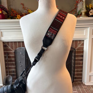 May include: A camera strap with a patterned design in red, black, and white is draped over a mannequin. The strap is attached to a black camera with a lens. The strap has a black leather backing and metal hardware.