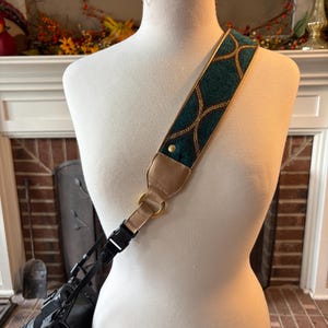 May include: A teal and gold camera strap with a decorative pattern is draped over a mannequin. The strap is attached to a black camera. The strap has a tan leather backing and gold hardware.