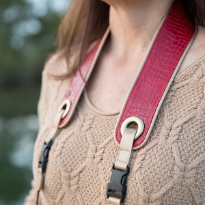 May include: A red camera strap with a crocodile-embossed leather design and gold-tone hardware. The strap is attached to a beige sweater.