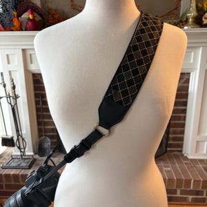 May include: A black camera with a patterned strap is displayed on a mannequin. The strap features a black and gold diamond pattern. The camera is attached to the strap with a black connector and gold ring.