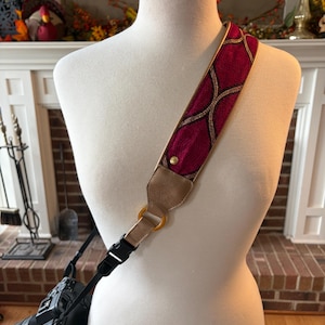May include: A camera strap with a burgundy and gold geometric design. The strap is attached to a black camera with a lens. The strap is displayed on a mannequin.