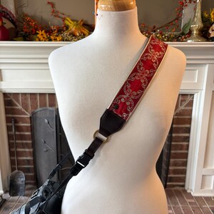 May include: A red and gold patterned camera strap is draped over a mannequin. The strap features a repeating floral design and is attached to a black camera. The background includes a fireplace and fall decorations.