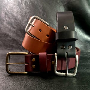 Puede incluir: Tres cinturones de cuero en tonos marrón, burdeos y negro. Cada cinturón presenta una hebilla metálica diferente: plateada, dorada y plateada. Los cinturones están apilados, mostrando sus diferentes colores y texturas.