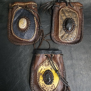 May include: Three rectangular leather pouches with drawstring closures. Each pouch features a central oval inlay with a textured pattern and a smooth, dark, polished stone. The edges are finished with braided leather stitching and metal eyelets.