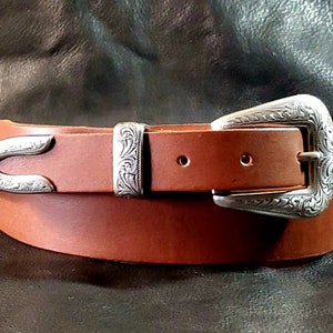 May include: A brown leather belt featuring a silver-toned metal buckle and a matching silver-toned metal belt tip and keeper. The metal components are decorated with an intricate, embossed floral scroll pattern throughout the surface.