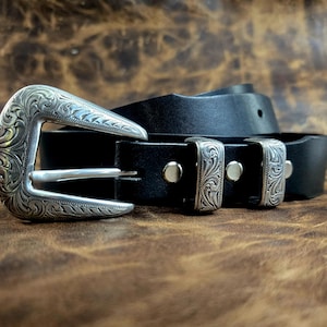 Peut inclure: Une ceinture en cuir noir dotée d'une grande boucle en métal argenté avec des gravures complexes en forme de volutes. La ceinture comprend deux passants en métal argenté assortis et des rivets argentés fixant la boucle à la sangle.
