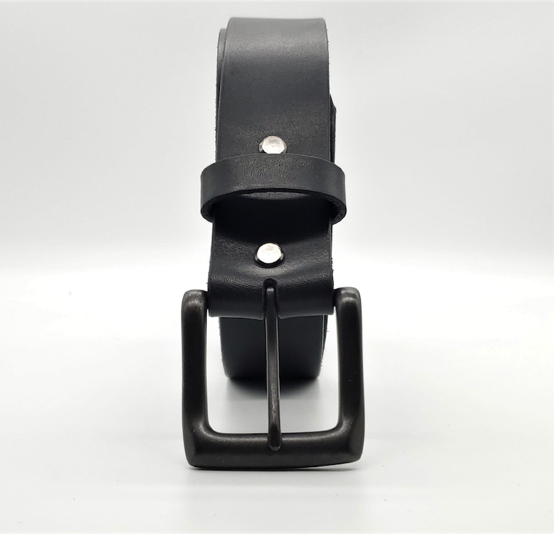 May include: A black leather belt with a dark metal buckle. The belt is rolled up, showing the buckle and the belt loop. The belt is secured with two silver rivets.