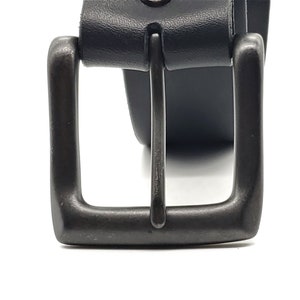 May include: Close-up of a black leather belt with a dark gray metal buckle. The buckle is square with a single metal bar. The leather belt is visible behind the buckle.