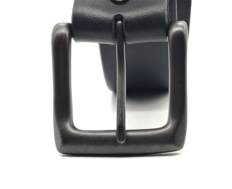 May include: Close-up of a black leather belt with a dark gray metal buckle. The buckle is square with a single metal bar. The leather belt is visible behind the buckle.