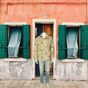 May include: A tan and brown camouflage jacket with multiple pockets and a collared neckline. The jacket is paired with light blue denim jeans. The mannequin is standing in front of a peach-colored building with green shutters and a wooden door.