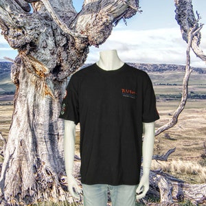 May include: Black t-shirt with the word "RUSH" in red script on the chest. The shirt has a small patch on the left sleeve and is displayed on a mannequin. The background features a landscape with a large tree.